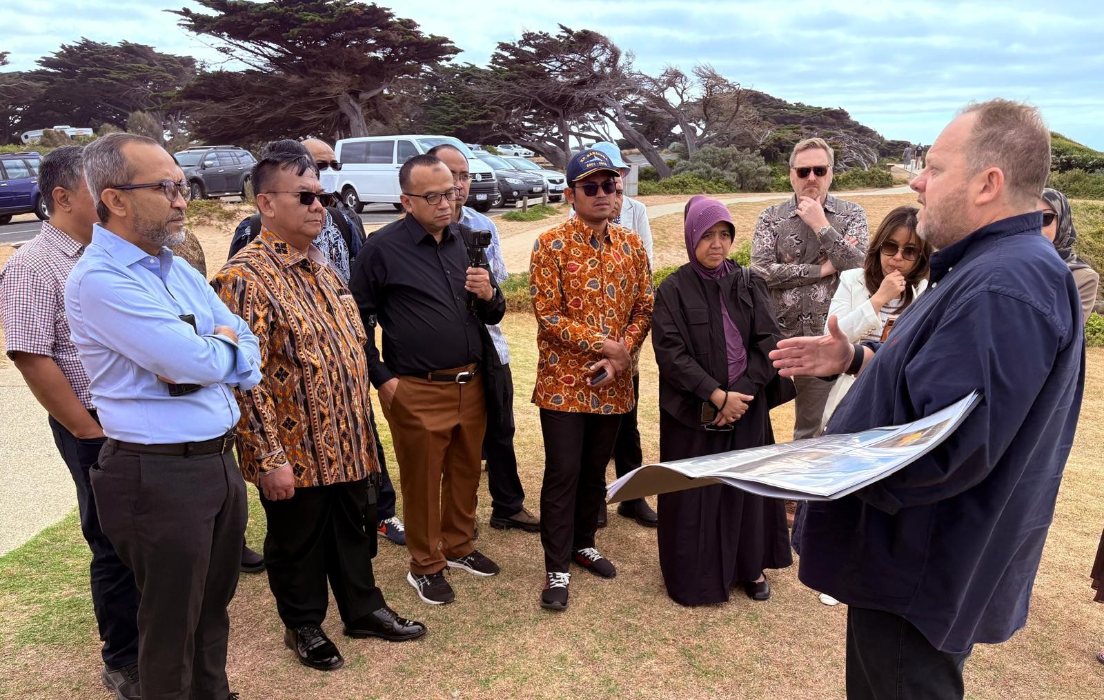 Representatives from the Government of Indonesia stand with Australian counterparts on a beach, one man talks to the group holding a informative picture as he talks about subsea telecommunication cables. 