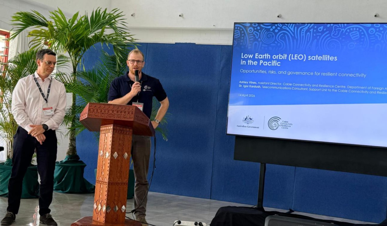 Two presenters speak at the front of a conference room, delivering a presentation on low Earth orbit satellite research.