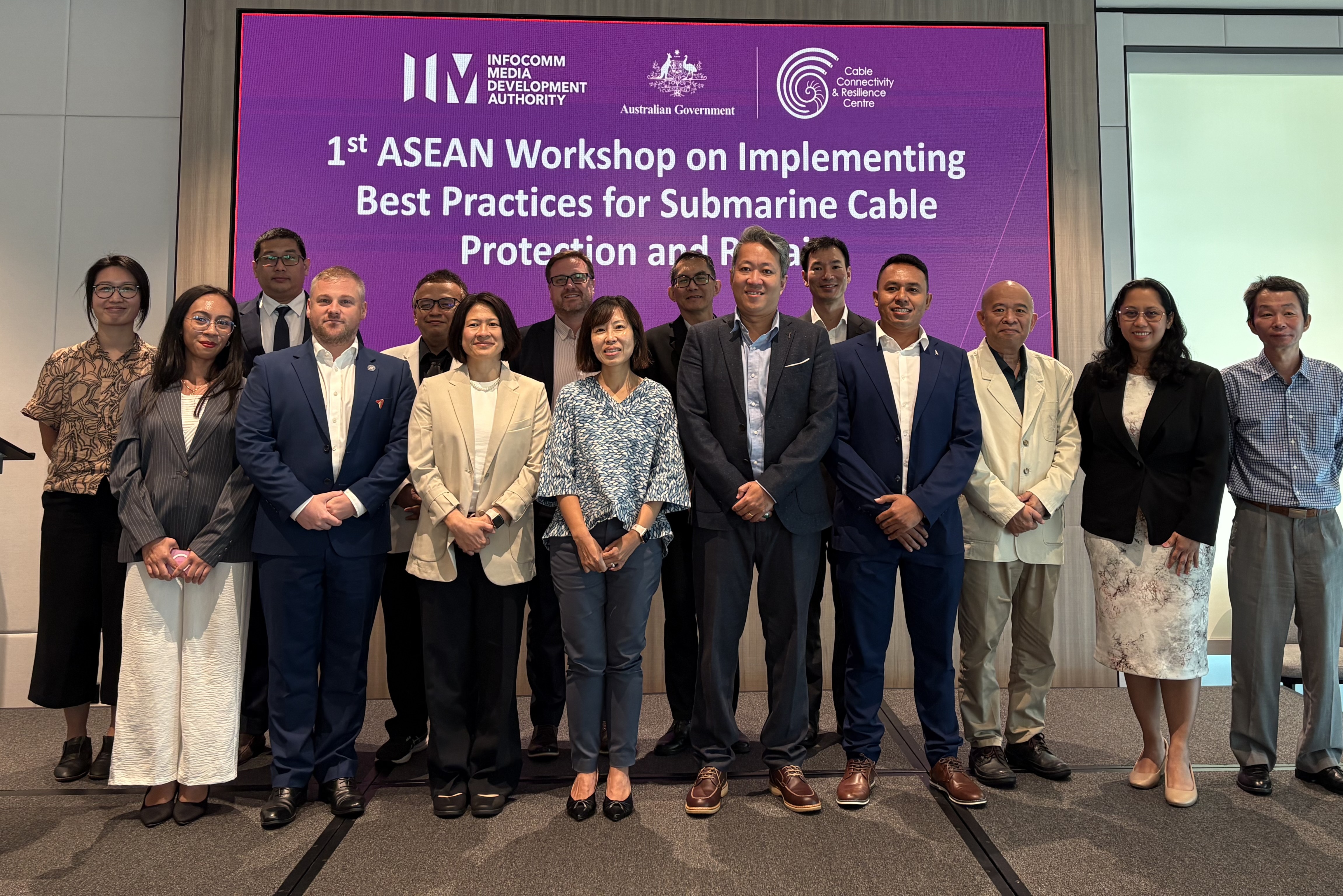 Workshop participants pose for a group photo in front of a screen with the name of the workshop, 1st ASEAN Workshop on Implementing Best Practices for Submarine Cable Protection and Repair. 