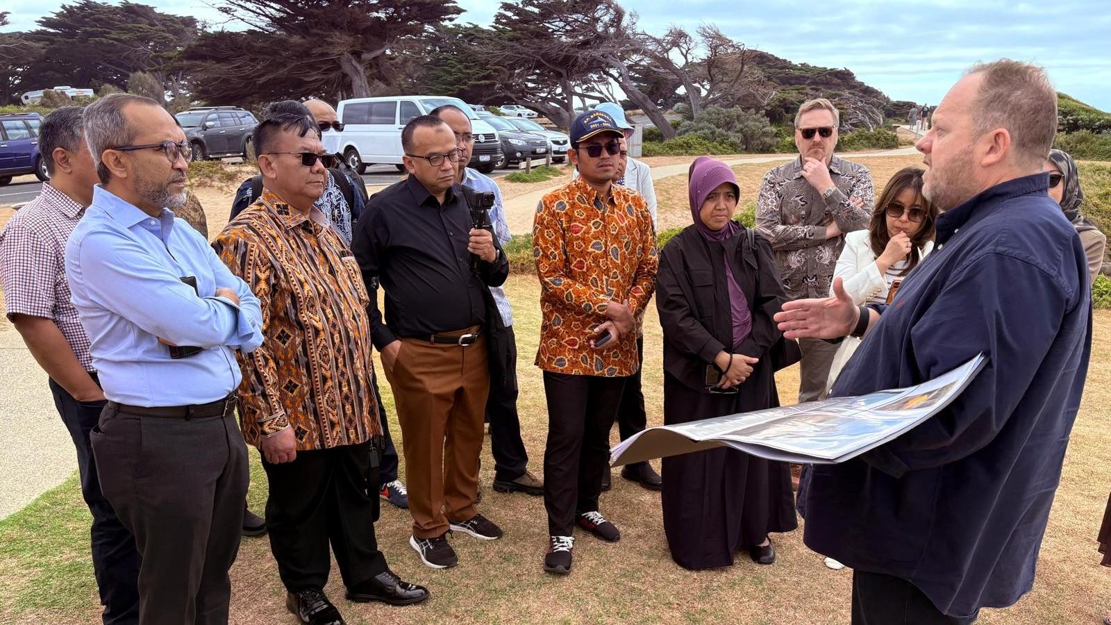 Representatives from the Government of Indonesia stand with Australian counterparts on a beach, one man talks to the group holding a informative picture as he talks about subsea telecommunication cables. 