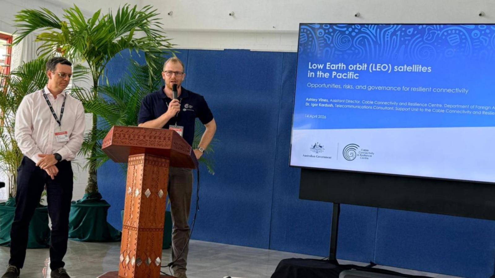 Two presenters speak at the front of a conference room, delivering a presentation on low Earth orbit satellite research.